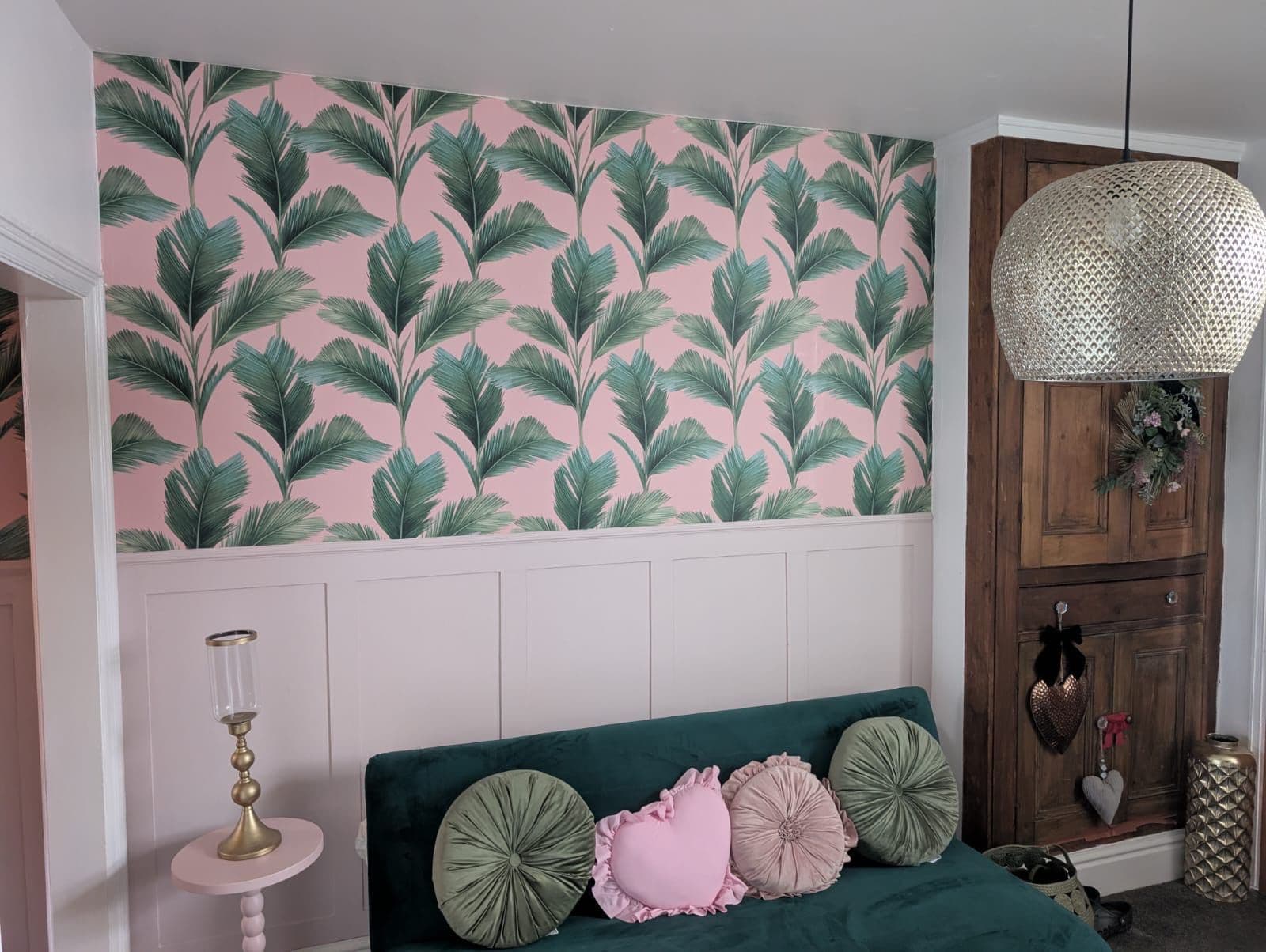 Room with pink palm leaf wallpaper above white shaker panelling and teal sofa